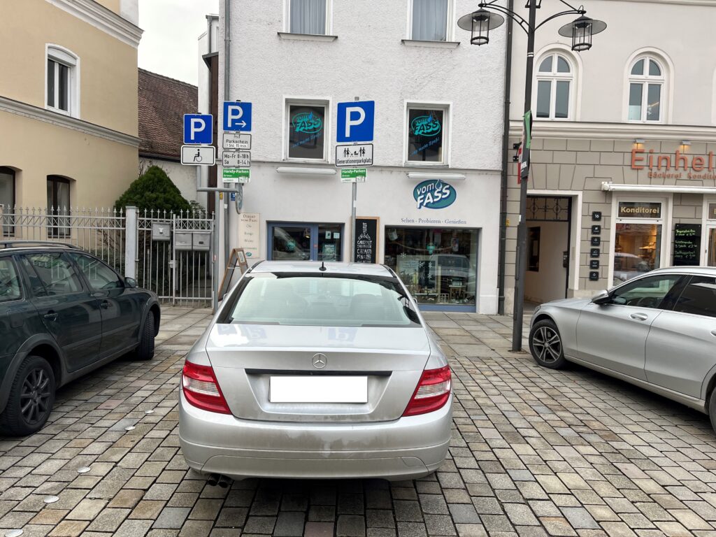 Ein silbernes Auto steht auf einem markierten Parkplatz vor einem Geschäft in einer gepflasterten Straße; daneben sind Park- und Verkehrsschilder sichtbar.