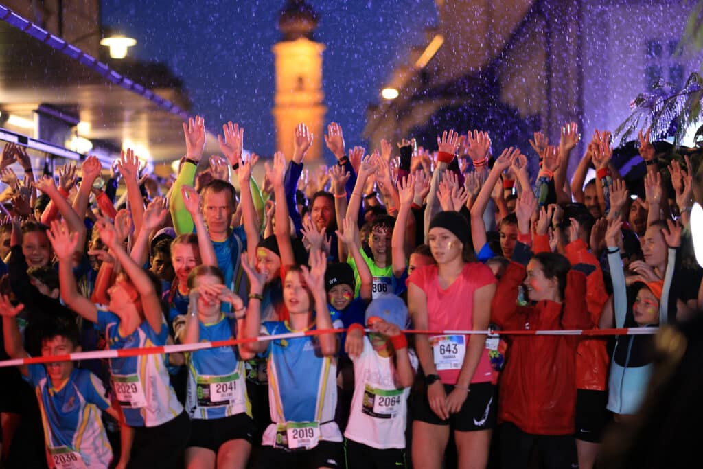 Startgruppe eines Nachtlaufs mit erhobenen Armen bei Regen vor beleuchteten Gebäuden
