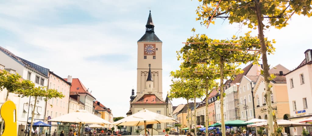 Blick über den Oberen Stadtplatz in Deggendorf mit der Stadtpfarrkirche, Cafés, Sonnenschirmen und herbstlich gefärbten Bäumen.