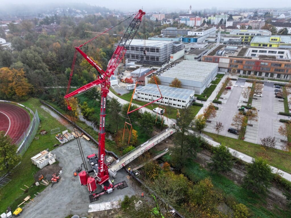 Neubau der Kollbachbrücke Roter Baukran auf einer Baustelle neben einer Sportanlage und Gewerbegebäuden; im Vordergrund wird eine Brücke über einen bewaldeten Einschnitt errichtet.