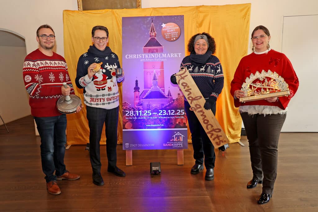 Fünf Menschen präsentieren ein Plakat zum Christkindlmarkt Deggendorf 2025; zwei tragen Weihnachtspullover, eine Person hält einen dekorierten Holzschlitten und eine weitere ein Tablett mit Gebäck.