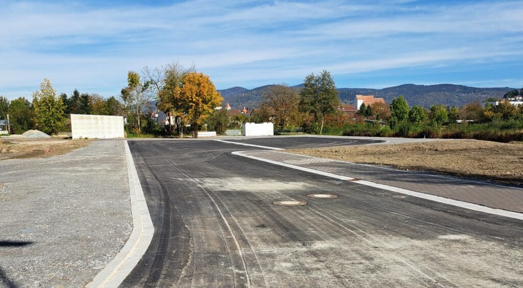 Neu angelegte Erschließungsstraße in den Schachinger Gärten mit Baumreihen, Häusern und bewaldeten Hügeln im Hintergrund.