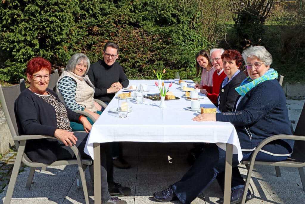 Senioren-Kaffeetrinken 2025 Acht ältere Menschen sitzen bei sonnigem Wetter an einer gedeckten Kaffeetafel im Garten.