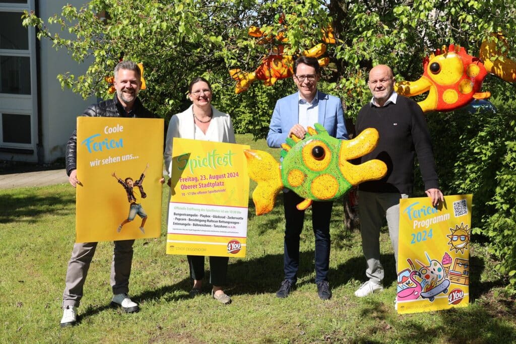Vier Personen präsentieren im Freien Plakate und eine große bunte Tierfigur zum 4You-Ferienprogramm; im Hintergrund hängt ein weiteres Ferienprogramm-Plakat.
