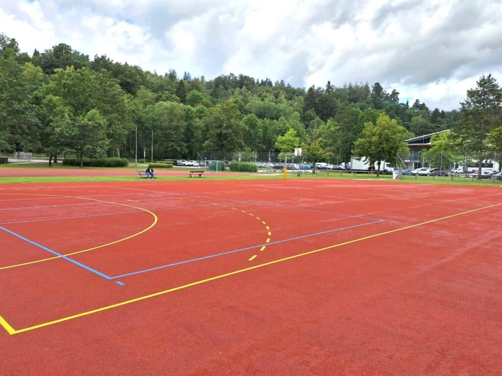 Sanierter Hartplatz im Berthold-Heckscher-Sportzentrum Roter, frisch sanierter Hartplatz mit gelben, blauen und weißen Spielfeldmarkierungen vor einem bewaldeten Hang.