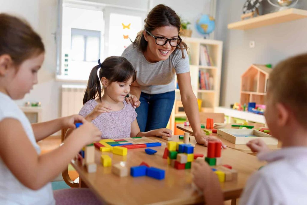 Eine lächelnde Erwachsene unterstützt drei Kinder beim Bauen mit bunten Holzklötzen an einem Tisch in einem hellen Klassenraum.