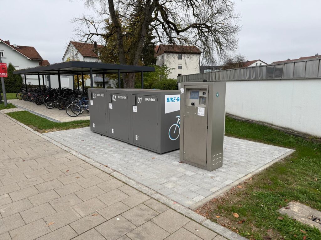 Überdachte Fahrradabstellanlage mit verschließbaren Fahrradboxen und einem Fahrradparkautomaten auf einem gepflasterten Platz.