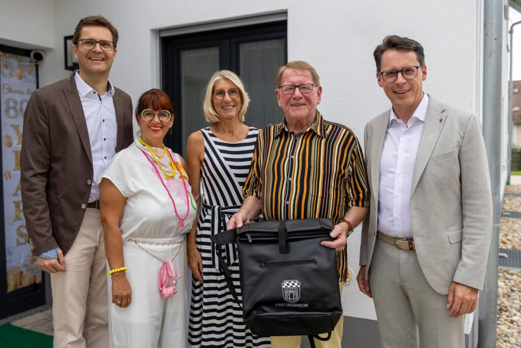Fünf Personen stehen vor einem Gebäude; in der Mitte hält ein Mann eine schwarze Tasche mit Wappen und Schriftzug.