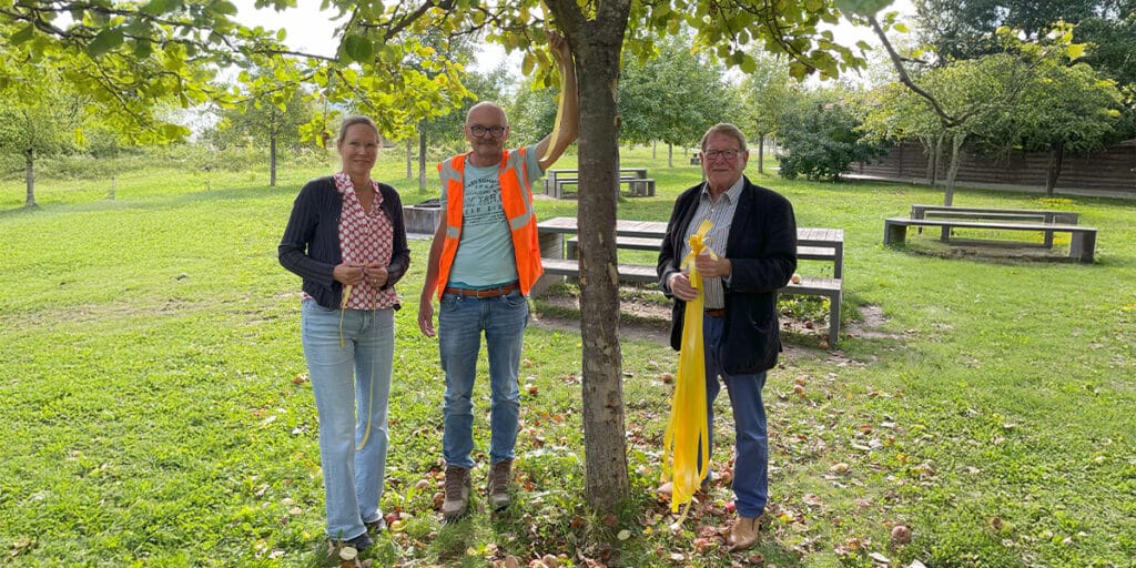 Drei Personen stehen auf einer Obstwiese neben einem Baum mit gelbem Band.
