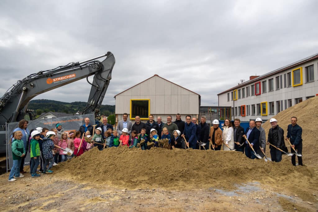 Gruppe aus Kindern und Erwachsenen steht mit Schaufeln vor einem Bagger und einem Gebäudekomplex auf einer Baustelle in Mietraching.