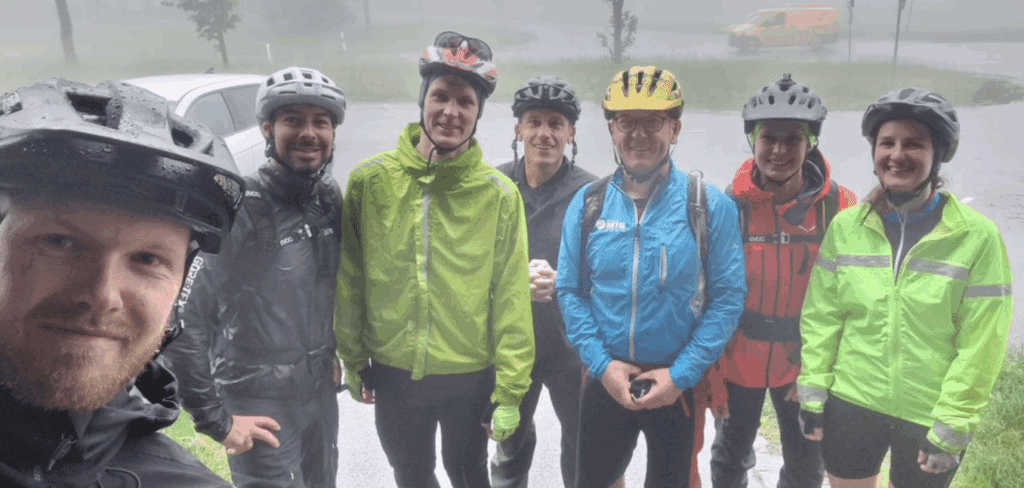 Sieben Radfahrende stehen bei Regen in Fahrradhelmen und wetterfester Kleidung für eine gemeinsame Aufnahme zusammen.