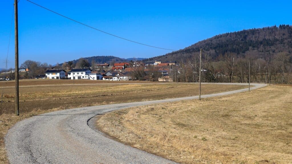 Kurvenreiche Schotterstraße durch eine offene Landschaft am Ledersberg; im Hintergrund liegen Häuser und ein bewaldeter Berghang.