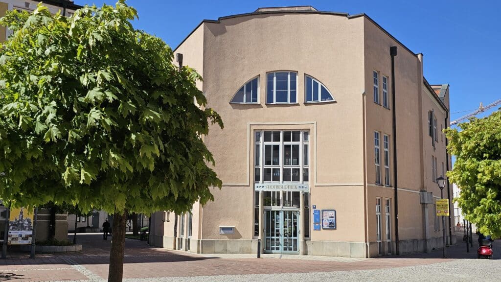Historisches, hellbraunes Gebäude mit hohen Fenstern und verglastem Eingangsbereich an einer begrünten Straße bei sonnigem Wetter.