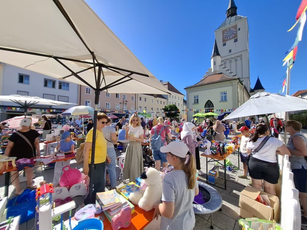 Belebter Kinderflohmarkt mit Verkaufsständen, Spielzeug und Besuchern auf einem Platz vor einer Kirche.