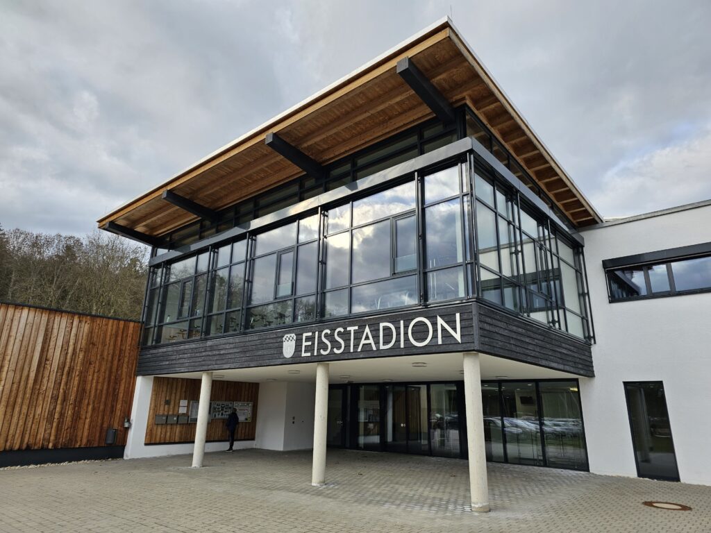 Eingangsbereich des Eisstadions mit verglaster Fassade, überdachtem Vorbau und dem Schriftzug „EISSTADION“.