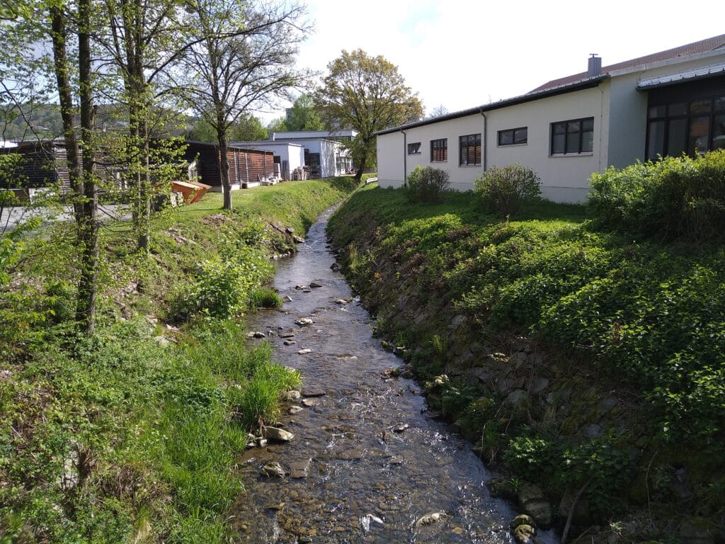 Der Hammermühlbach fließt zwischen begrünten Ufern an einem Gebäude entlang.