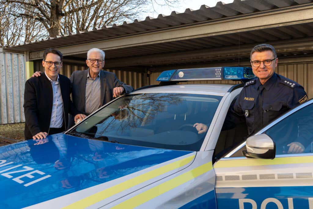 Neujahrskonzert 2026 in Deggendorf Drei Männer stehen neben einem blau-silbernen Polizeifahrzeug mit eingeschaltetem Blaulicht vor einem Gebäude.