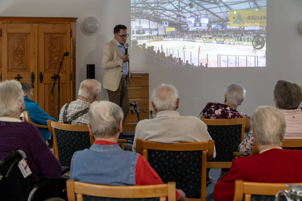 Ältere Menschen sitzen bei einer Präsentation im Haus der Diakonie; an der Wand ist eine Eishockeyarena zu sehen.