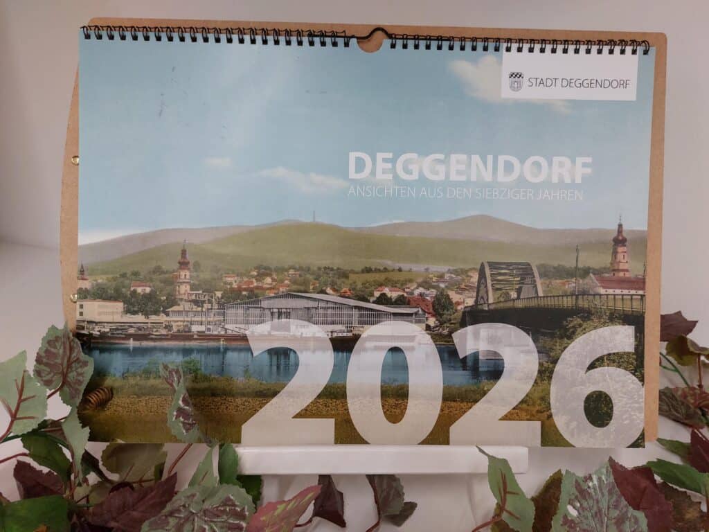 Aufgeschlagener Fotokalender 2026 mit einer Stadtansicht von Deggendorf an der Donau, dekoriert mit künstlichen Blättern.
