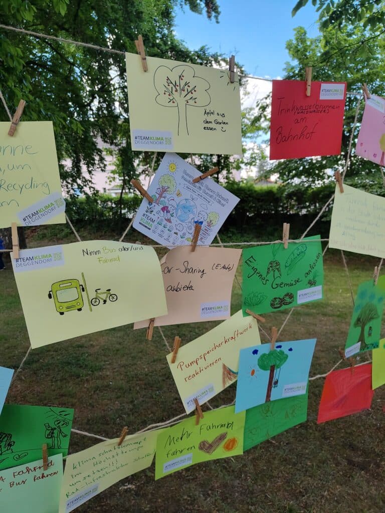 Bunte handgeschriebene Plakate mit Umwelt- und Nachhaltigkeitsmotiven hängen zwischen Bäumen im Freien.