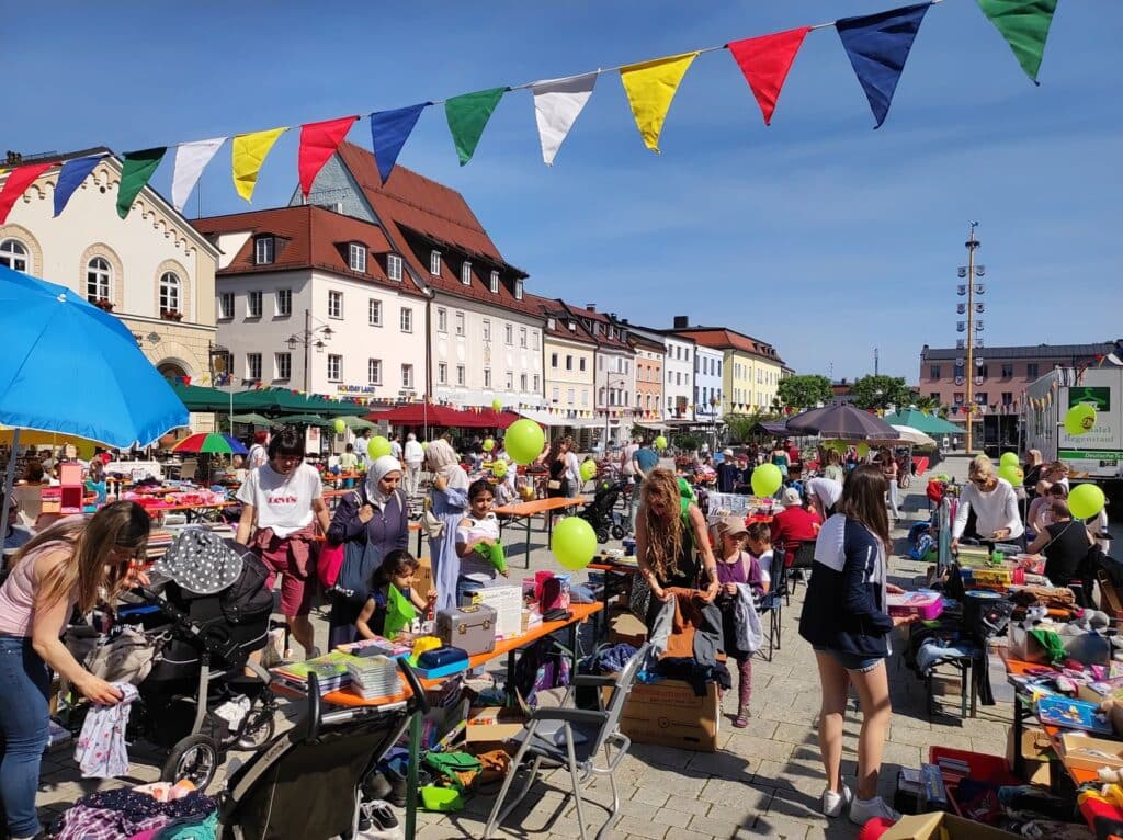 Belebter Kinderflohmarkt auf einem Stadtplatz mit Verkaufsständen, Besuchenden, Kinderwagen, bunten Luftballons und Wimpelketten