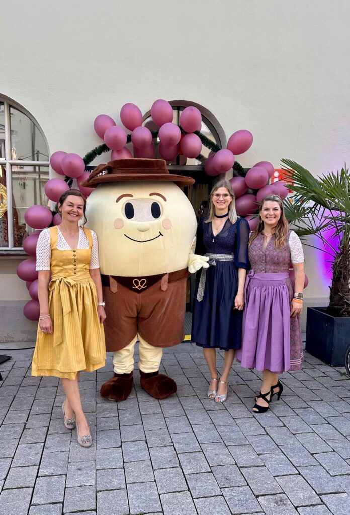 Ein großes lächelndes Maskottchen mit violetten Luftballons steht neben drei Frauen in Dirndln vor einem Gebäude.