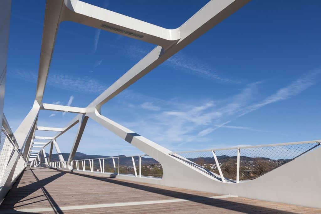 Weiße Fußgängerbrücke mit markanter Stahlkonstruktion und Holzbelag vor blauem Himmel und bewaldeten Hügeln.