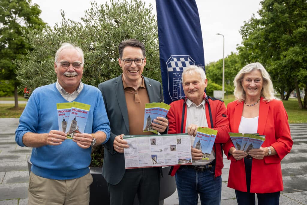 Neues Seniorenprogramm in Deggendorf vorgestellt Vier lächelnde Erwachsene präsentieren Broschüren und ein aufgefaltetes Programmheft vor einem blauen Banner im Freien.