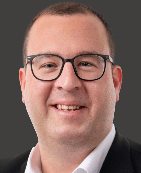 Porträt eines lächelnden Mannes mit schwarzer Brille und dunklem Anzug vor grauem Hintergrund
