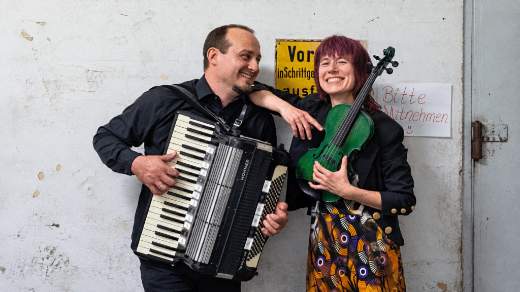 Zwei lächelnde Musikerinnen und Musiker vor einer Wand mit Akkordeon und grünem Saiteninstrument