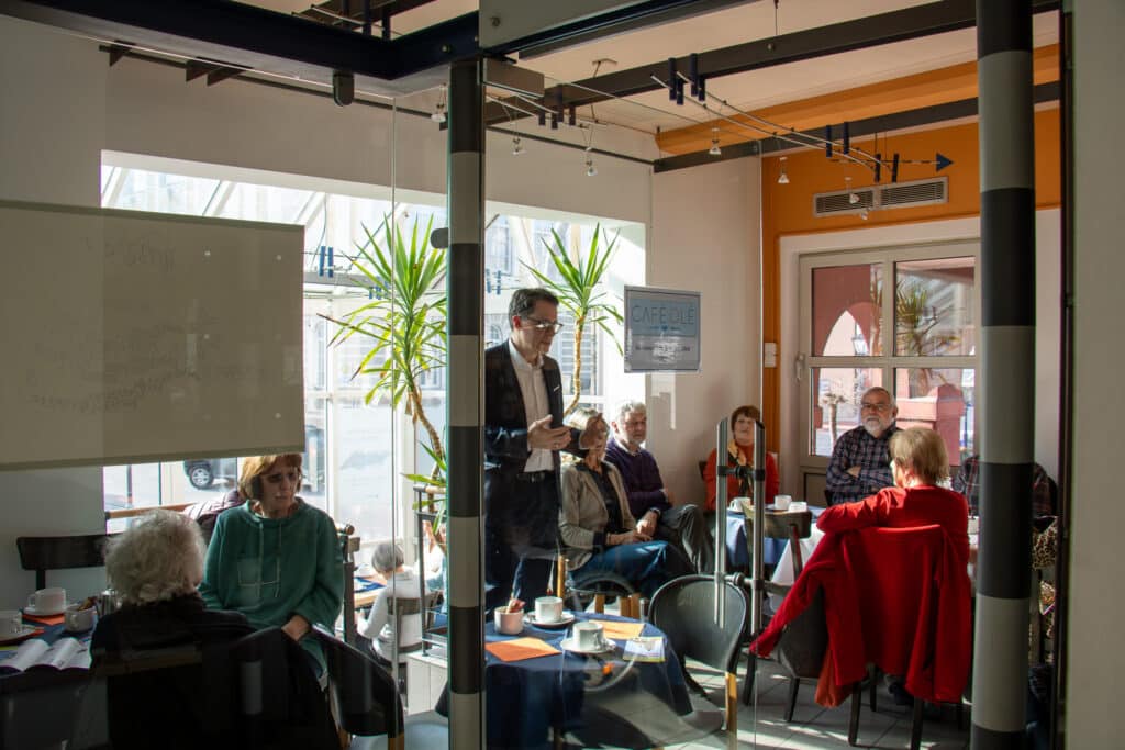 Mehrere Menschen sitzen bei einem Treffen an Tischen in einem hellen Café- oder Veranstaltungsraum; eine Person steht in der Mitte und spricht.