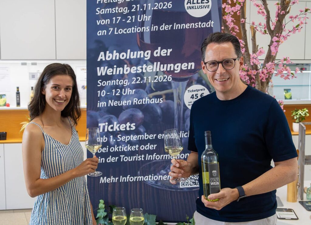 Zwei Personen präsentieren Weinflaschen vor einem Veranstaltungsplakat zu „Weinloben in Deggendorf“ im Alten Rathaus.