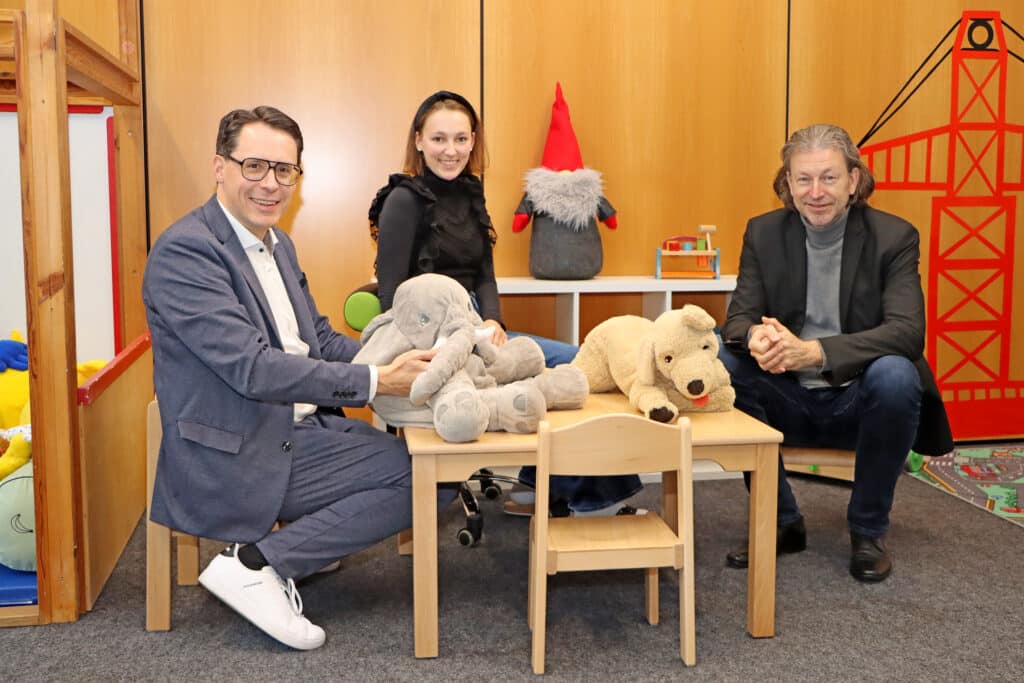 Vorstellung der Erweiterung des Kindergartens Aman Drei Erwachsene sitzen in einem Kindergartenraum an einem Tisch mit Stofftieren und Weihnachtsdekoration.