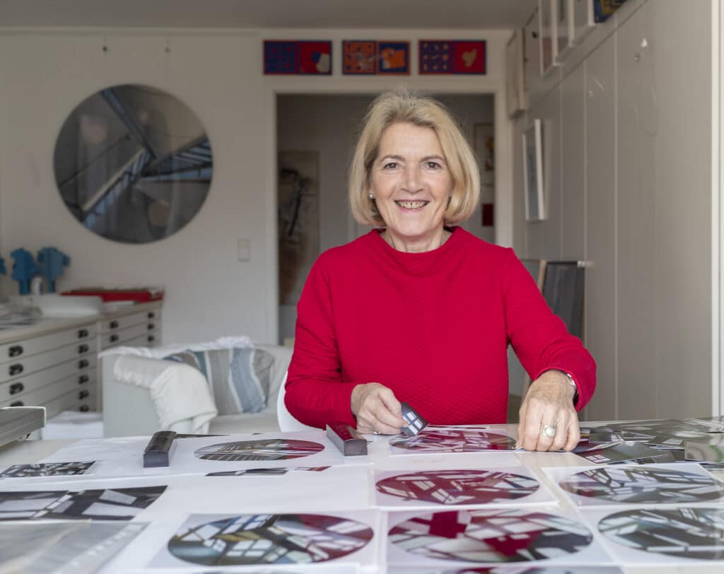 Lächelnde Frau mit kurzen blonden Haaren sitzt in einem roten Pullover an einem Tisch mit ausgebreiteten Fotografien und Unterlagen.