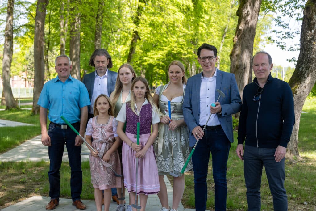 Acht Personen stehen mit Minigolfschlägern auf einer Minigolfanlage im grünen Stadtpark.