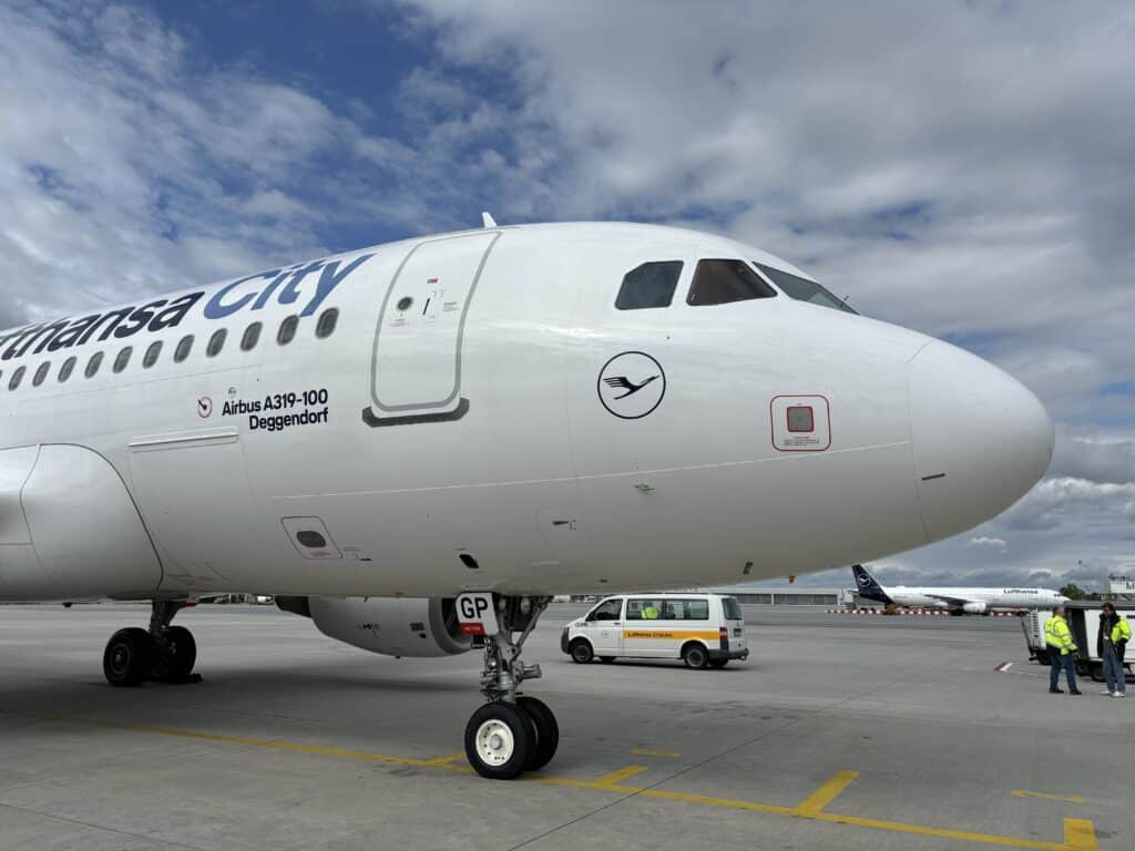 Nahaufnahme eines weißen Airbus A319-100 mit der Aufschrift „Deggendorf“ auf dem Rollfeld; im Hintergrund stehen ein Servicefahrzeug und weitere Flugzeuge.