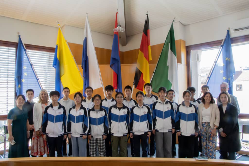 Gruppe chinesischer Austauschschülerinnen und Austauschschüler mit Begleitpersonen steht im Rathaus vor mehreren National- und Europaflaggen.