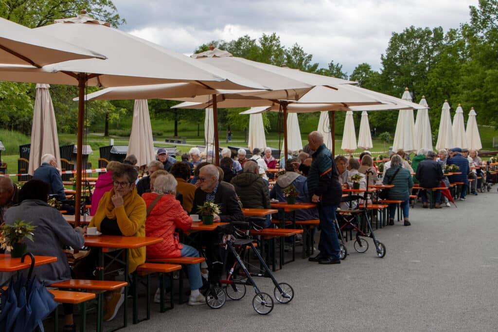 Seniorinnen und Senioren sitzen bei einer geselligen Kaffeerunde unter Sonnenschirmen im Außenbereich eines Cafés.