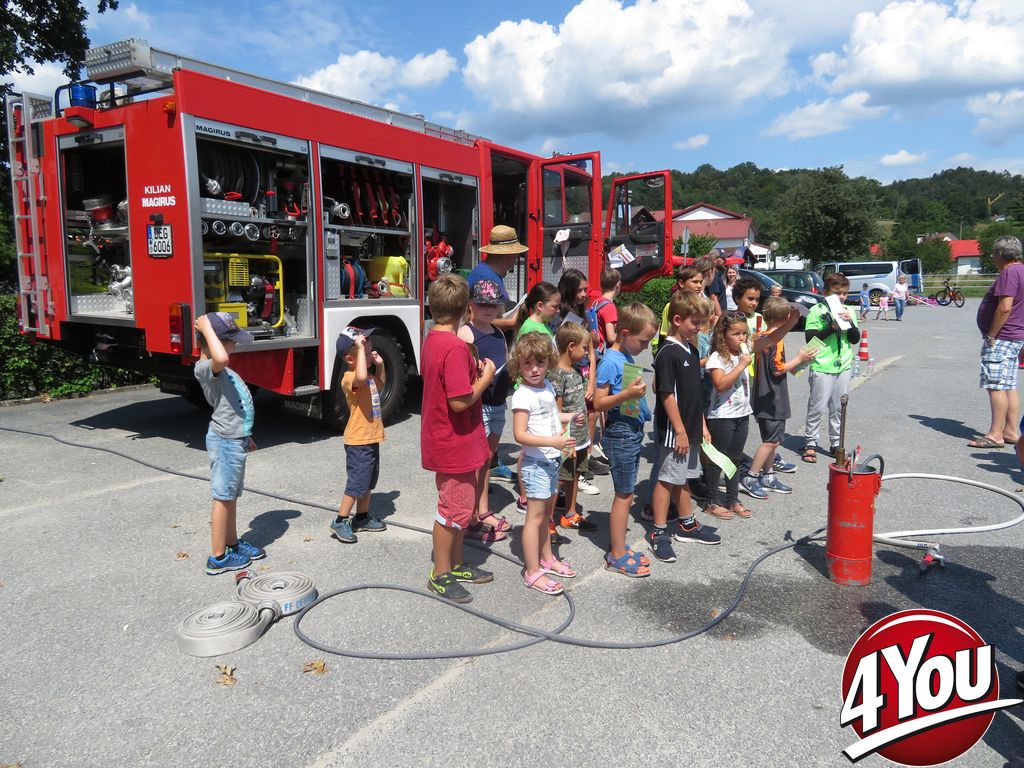 Kinder und Jugendliche stehen neben einem roten Feuerwehrfahrzeug und beobachten eine Vorführung mit einem roten Feuerlöscher.