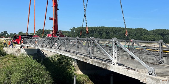 Brücke während Bauarbeiten Temporäre Stahlbrücke über einem bewachsenen Geländeeinschnitt; an der Brücke stehen ein roter Kran und mehrere Menschen.
