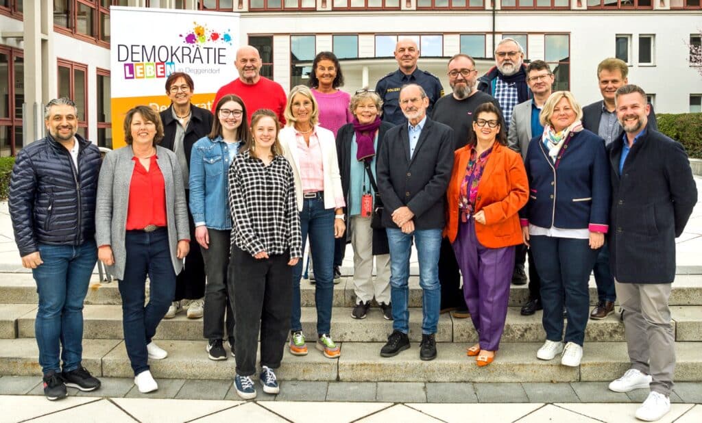 Gruppenfoto von 17 Bündnismitgliedern auf den Stufen vor einem Rathaus; im Hintergrund ist ein Schild mit dem Schriftzug „Demokratie leben!“ zu sehen.