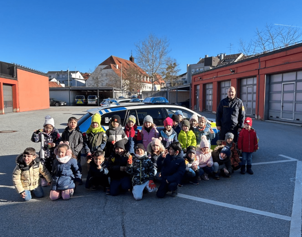 Gruppe von Vorschulkindern mit einer erwachsenen Begleitperson auf dem Hof eines Polizeigebäudes; im Hintergrund sind Polizeifahrzeuge zu sehen.