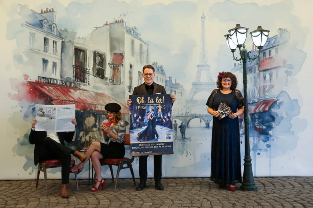 Vier Personen vor einer gemalten Pariser Straßenszene mit Eiffelturm; eine Person hält ein Plakat zur Veranstaltung „Le Bal de Paris“, zwei weitere sitzen mit Zeitungen auf Stühlen.