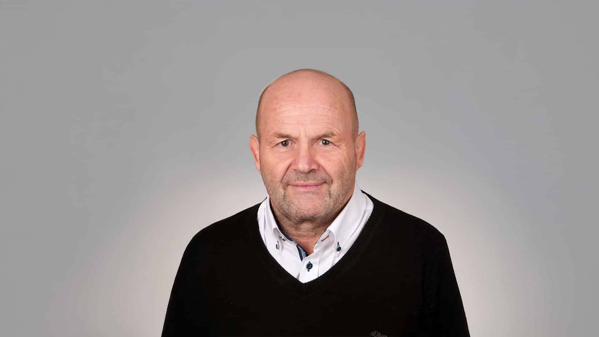 Porträt eines lächelnden, kahlköpfigen Mannes mit schwarzem Pullover und weißem Hemd vor grauem Hintergrund.