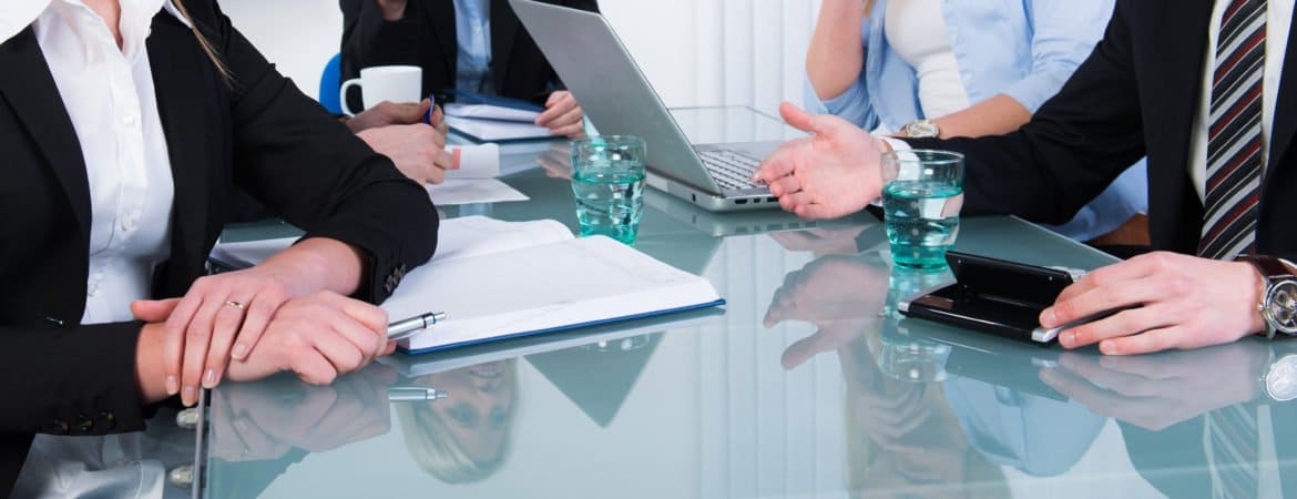Mehrere Personen besprechen sich an einem Glastisch; auf dem Tisch liegen Unterlagen, Laptops und Getränke.