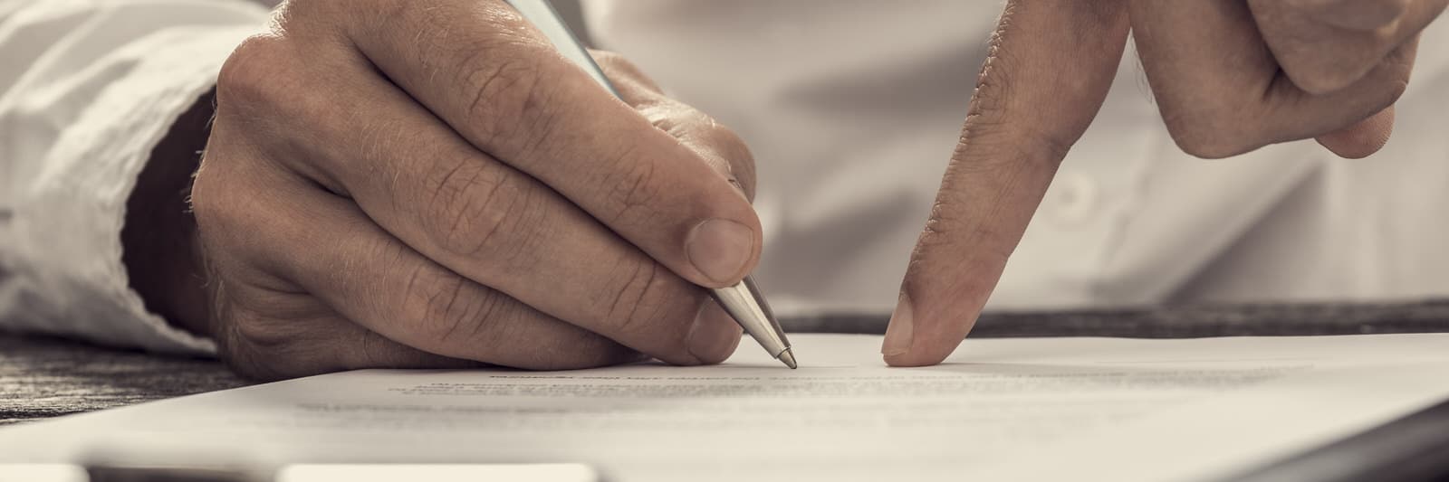 Nahaufnahme von zwei Händen über einem Dokument: Eine Hand schreibt mit einem Stift, während die andere auf eine Stelle im Papier zeigt.
