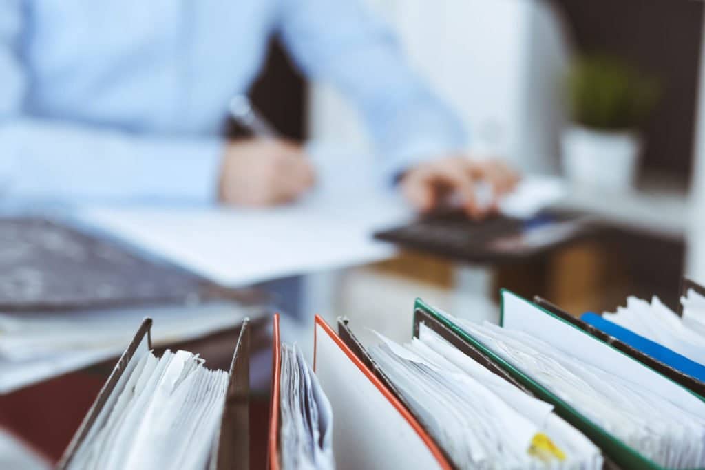 Geöffnete Aktenordner und Verwaltungsunterlagen im Vordergrund, dahinter eine unscharfe Person an einem Schreibtisch