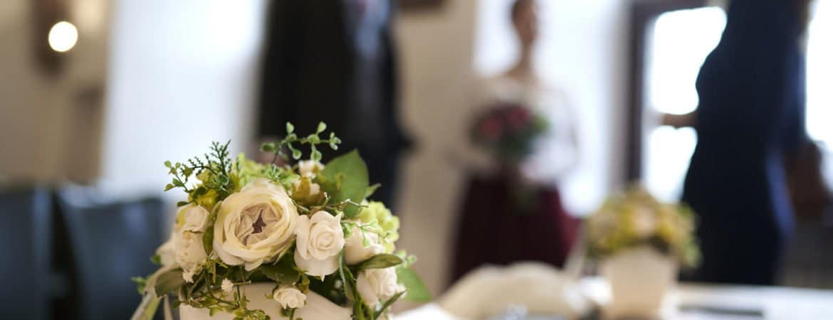 Standesamtliche Trauung im Deggendorfer Rathaus Blumenarrangement auf einem Tisch im Trauzimmer; im Hintergrund stehen mehrere unscharfe Personen.
